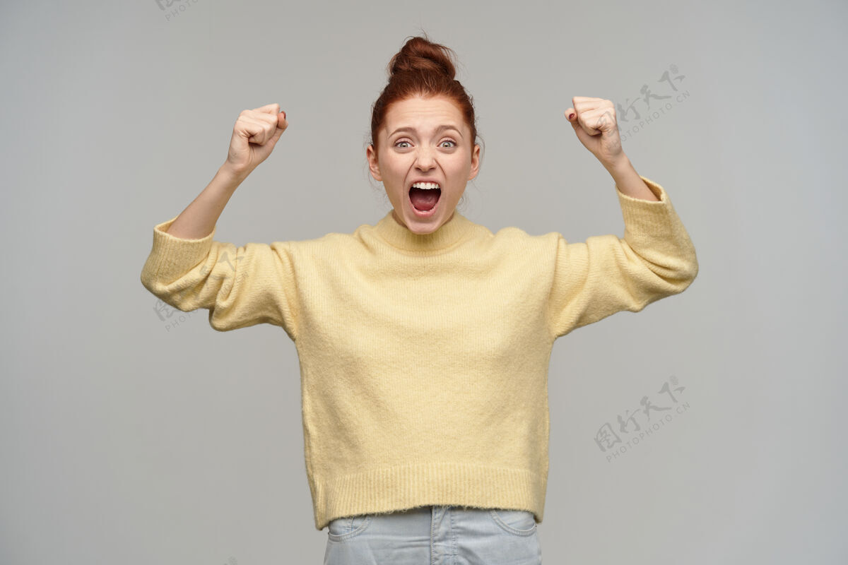 情感兴奋的红发女孩的画像 头发盘成发髻 穿着淡黄色毛衣和牛仔裤 举起拳头大喊 庆祝胜利隔着灰墙孤立女士女孩表情