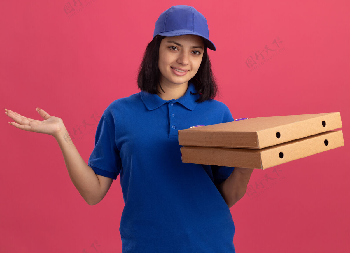 站着身穿蓝色制服 头戴鸭舌帽 手持披萨盒的年轻送货女孩友好地微笑着 举起手臂站在粉红色的墙上送货微笑披萨