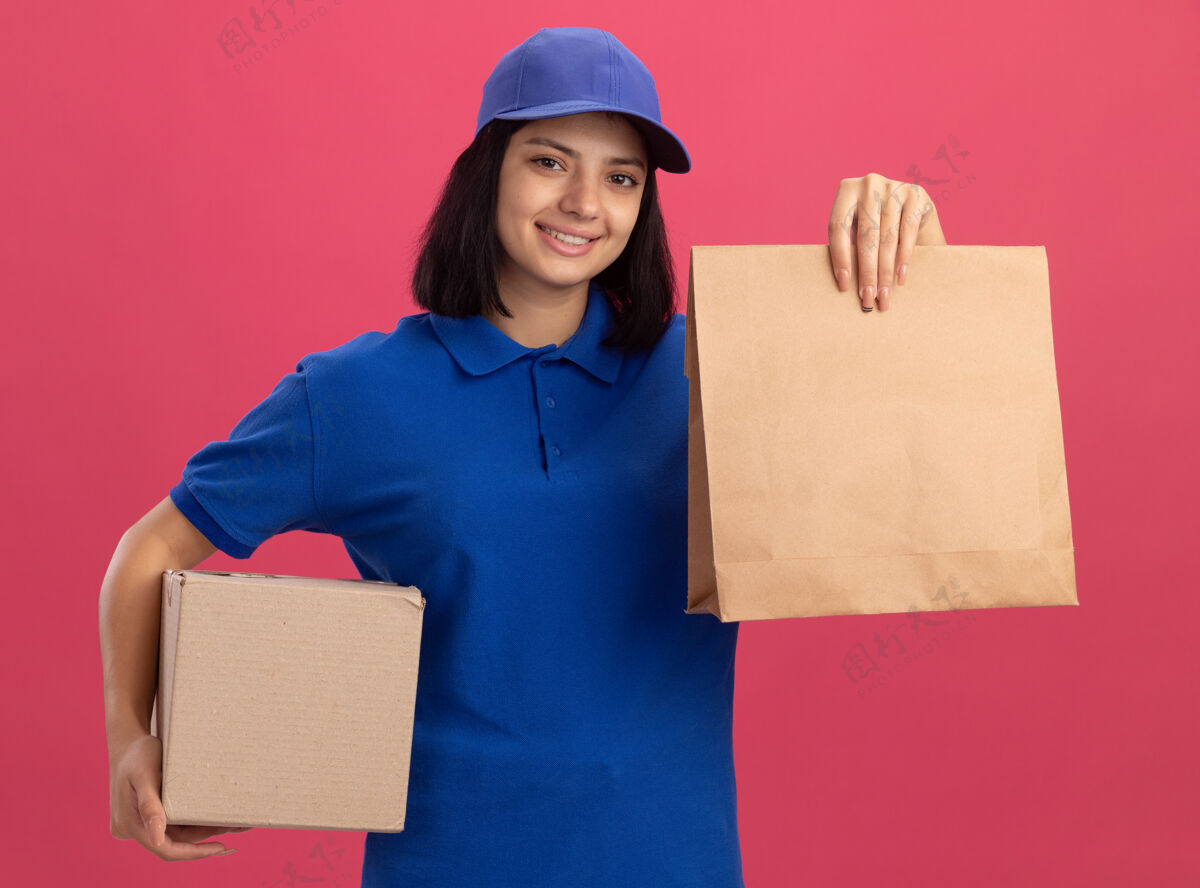 站着身穿蓝色制服 头戴鸭舌帽 手持纸包装和纸板箱的年轻送货女孩站在粉红色的墙上 脸上露出幸福的笑容年轻送货微笑