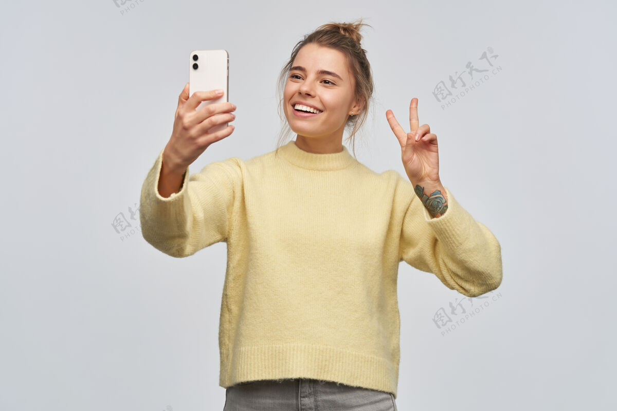 20多岁一个漂亮的成年女孩的肖像 金发束成发髻 纹身穿着黄色毛衣 手持智能手机自拍显示和平标志隔着白墙孤立地站着女士模特纹身