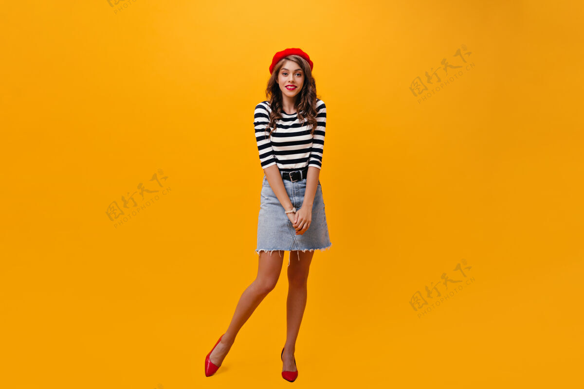 年轻身着牛仔裙 头戴红色贝雷帽的女士在橙色背景下摆出姿势穿着时髦服装和明亮高跟鞋的年轻女子正对着镜头女人空间复制