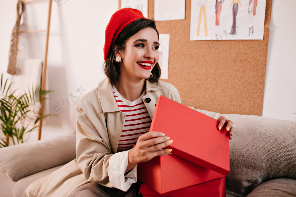 休闲穿米色风衣的黑发女人微笑着打开红色的盒子穿着条纹毛衣和鲜艳帽子的漂亮可爱的女孩微笑着红唇礼品盒公寓
