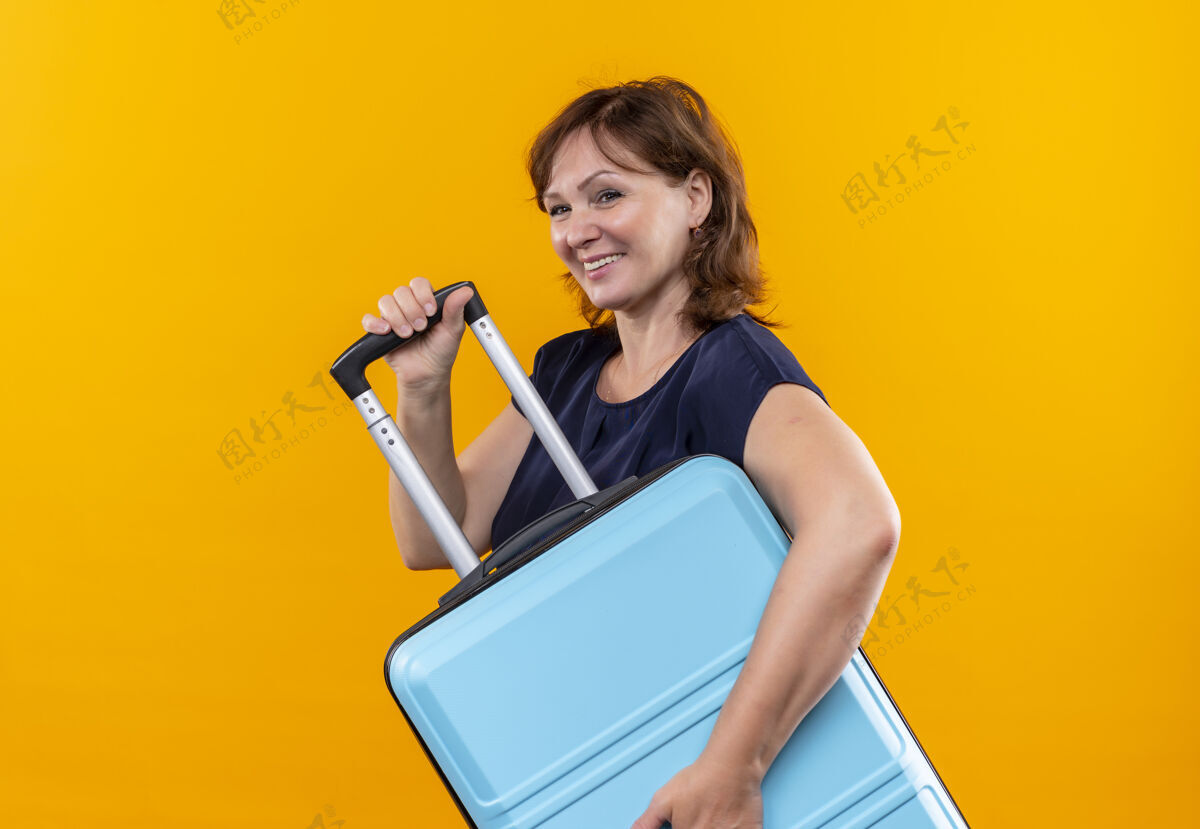抱着看着身边微笑的中年旅行家妇女 手拿手提箱 背景是孤立的黄色微笑壁板手提箱