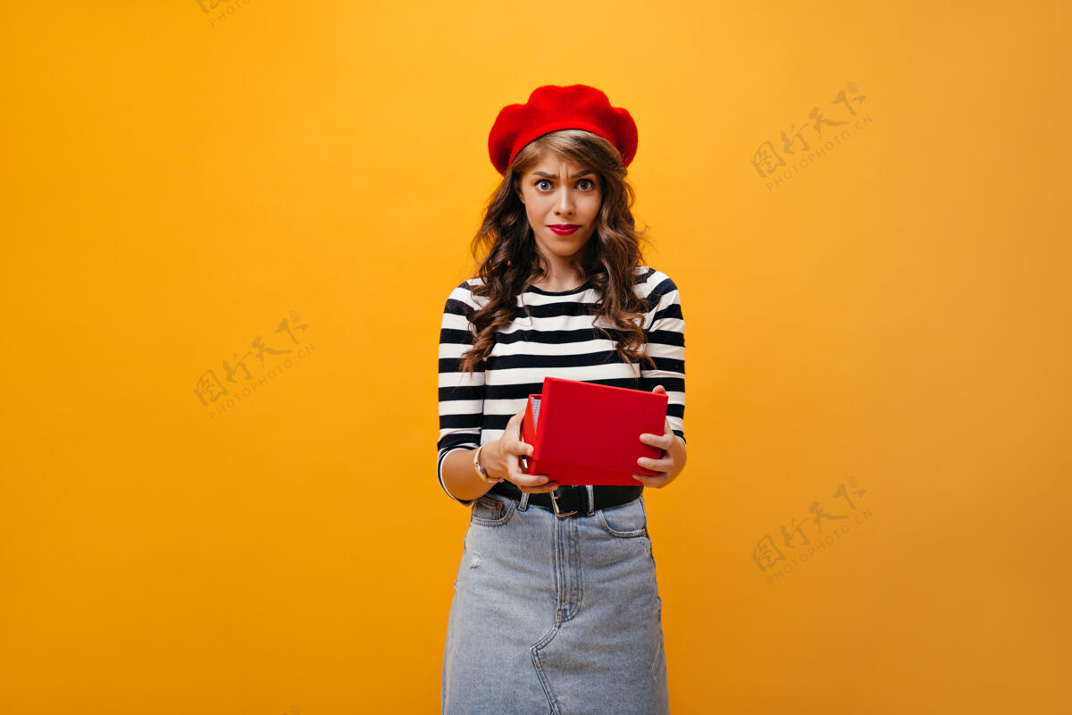 衬衫打开红色盒子后 女人看起来很不满意悲伤的女孩卷发 戴红色贝雷帽 牛仔裙 腰带 在孤立的背景下摆姿势礼物女性一个