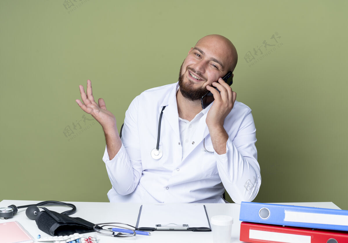 请高兴的年轻秃头男医生穿着医用长袍和听诊器坐在办公桌前 拿着医疗工具 在绿色背景下讲电话坐着穿电话