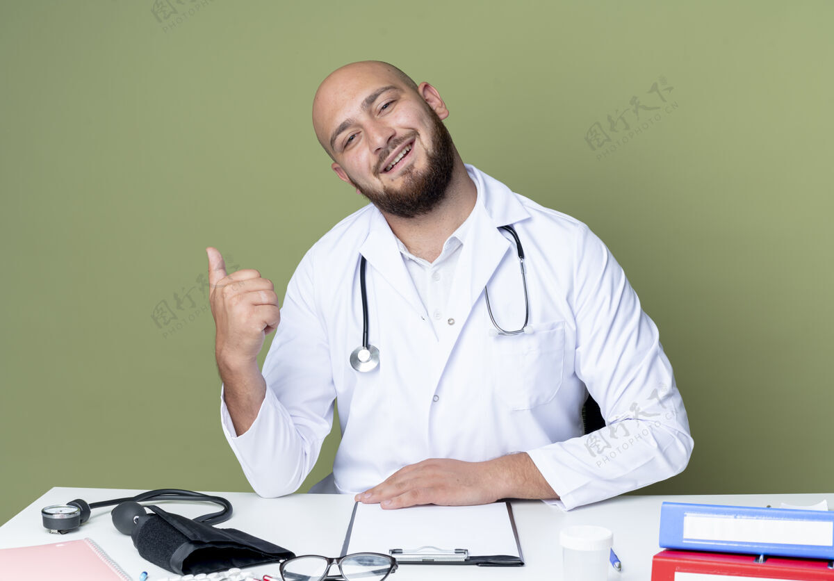 工具高兴的年轻秃头男医生穿着医用长袍和听诊器坐在办公桌前 用医用工具点在一旁 隔离在绿色背景上长袍点办公桌