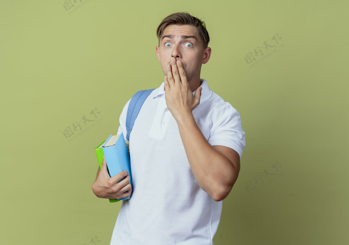 男性看着镜头吓得年轻帅气的男学生背着书包 捂着嘴 手孤立在橄榄绿的背景上穿害怕嘴