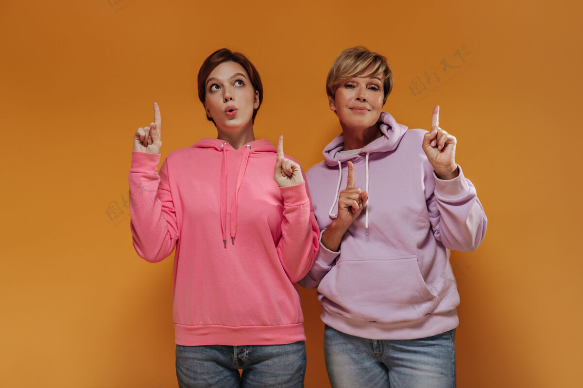 成熟两个酷酷的女人 短发 粉色和淡紫色宽连帽衫 牛仔裤 竖起大拇指 摆出橙色背景的姿势女儿年龄连帽衫
