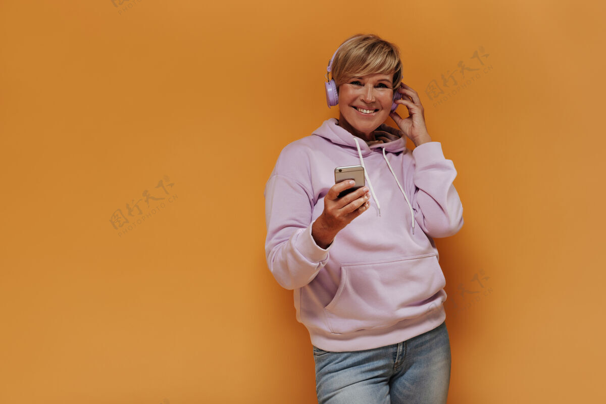 成熟快乐的老妇人 时髦的金发 淡紫色的耳机 时髦的粉色连帽衫和牛仔裤 微笑着拿着智能手机老年人橙色背景肖像