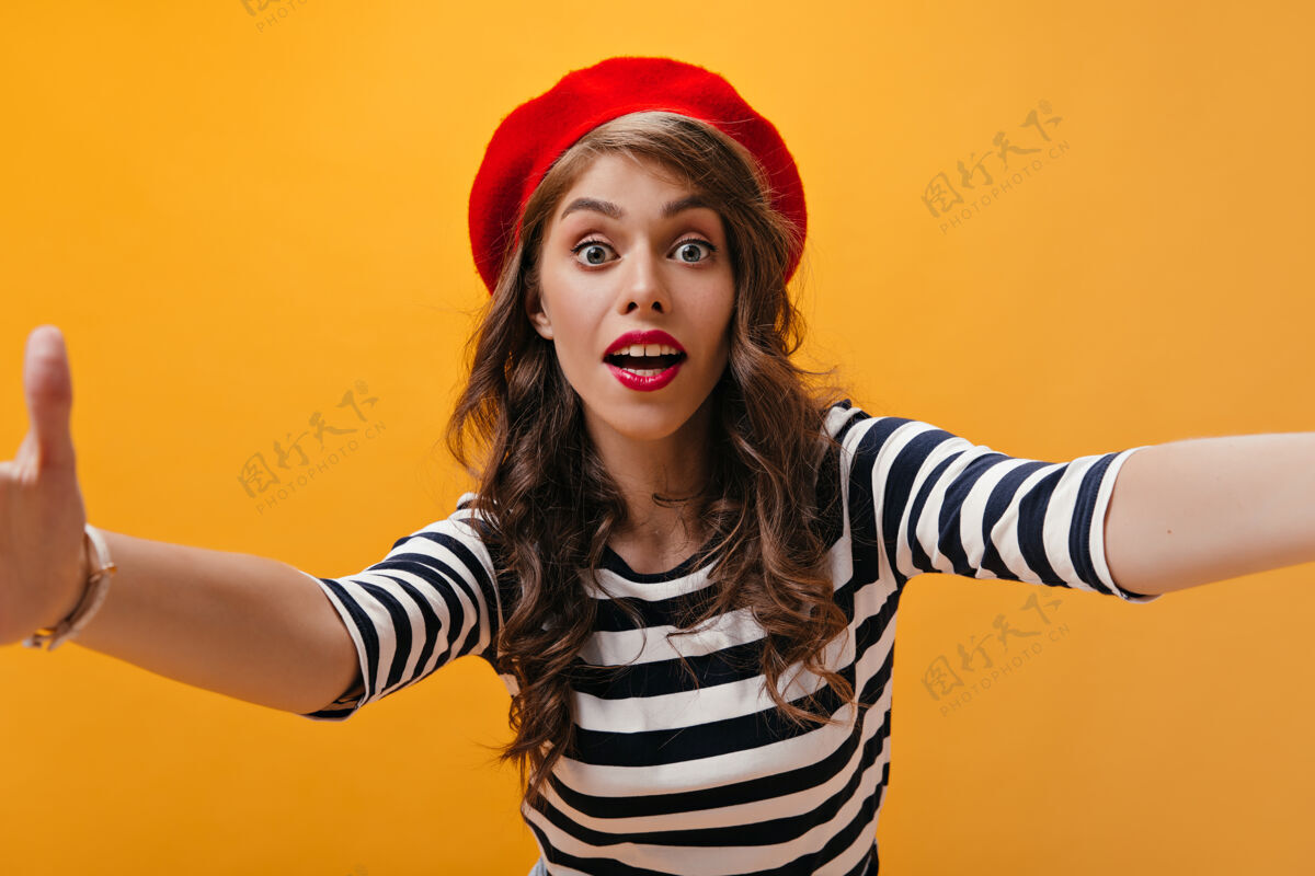 帽子身着红色贝雷帽的惊艳女士在橙色背景上自拍卷发的现代女性 穿着条纹衣服摆出明亮的嘴唇条纹年轻头发