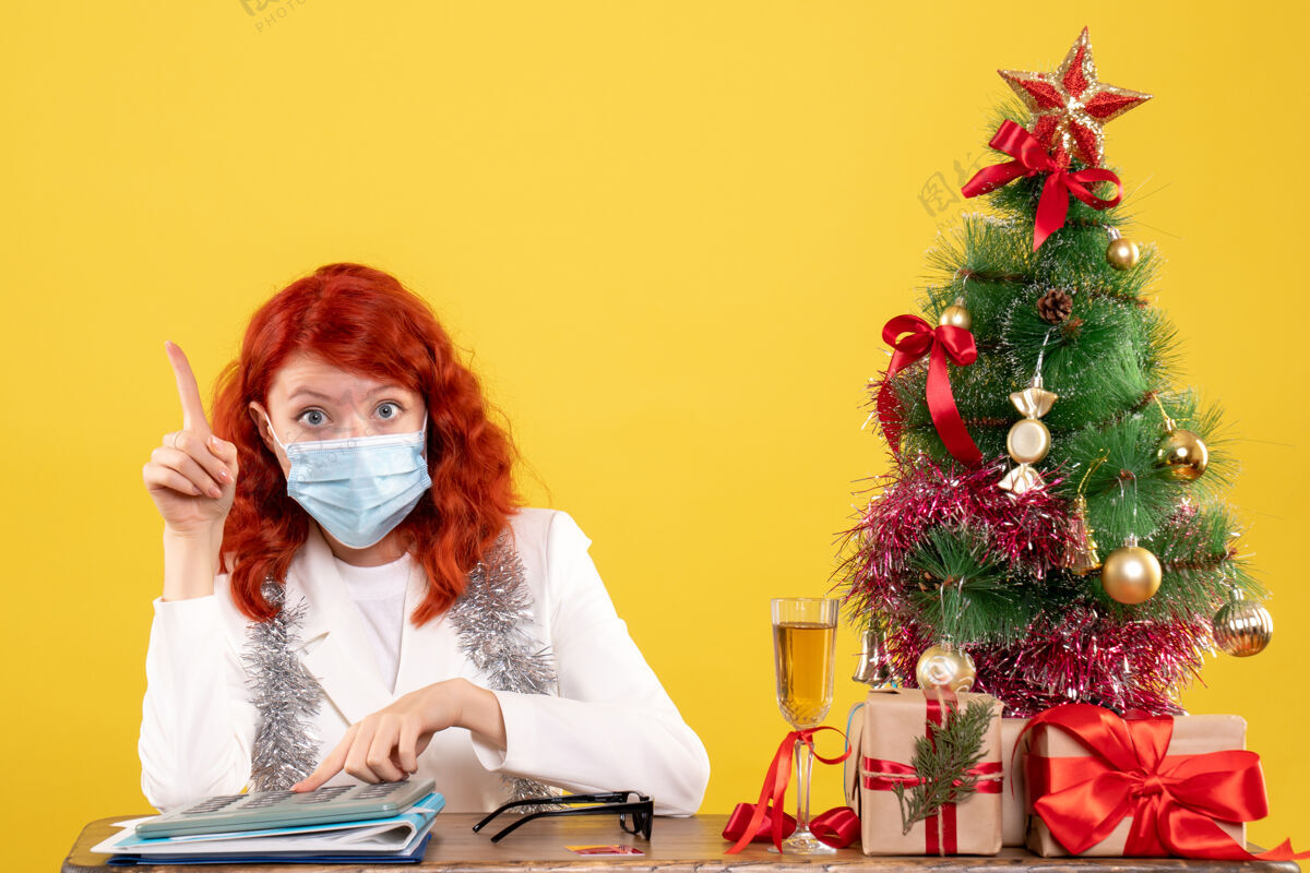 面具前视图女医生围着圣诞树 戴着消毒口罩坐着成人礼物美丽