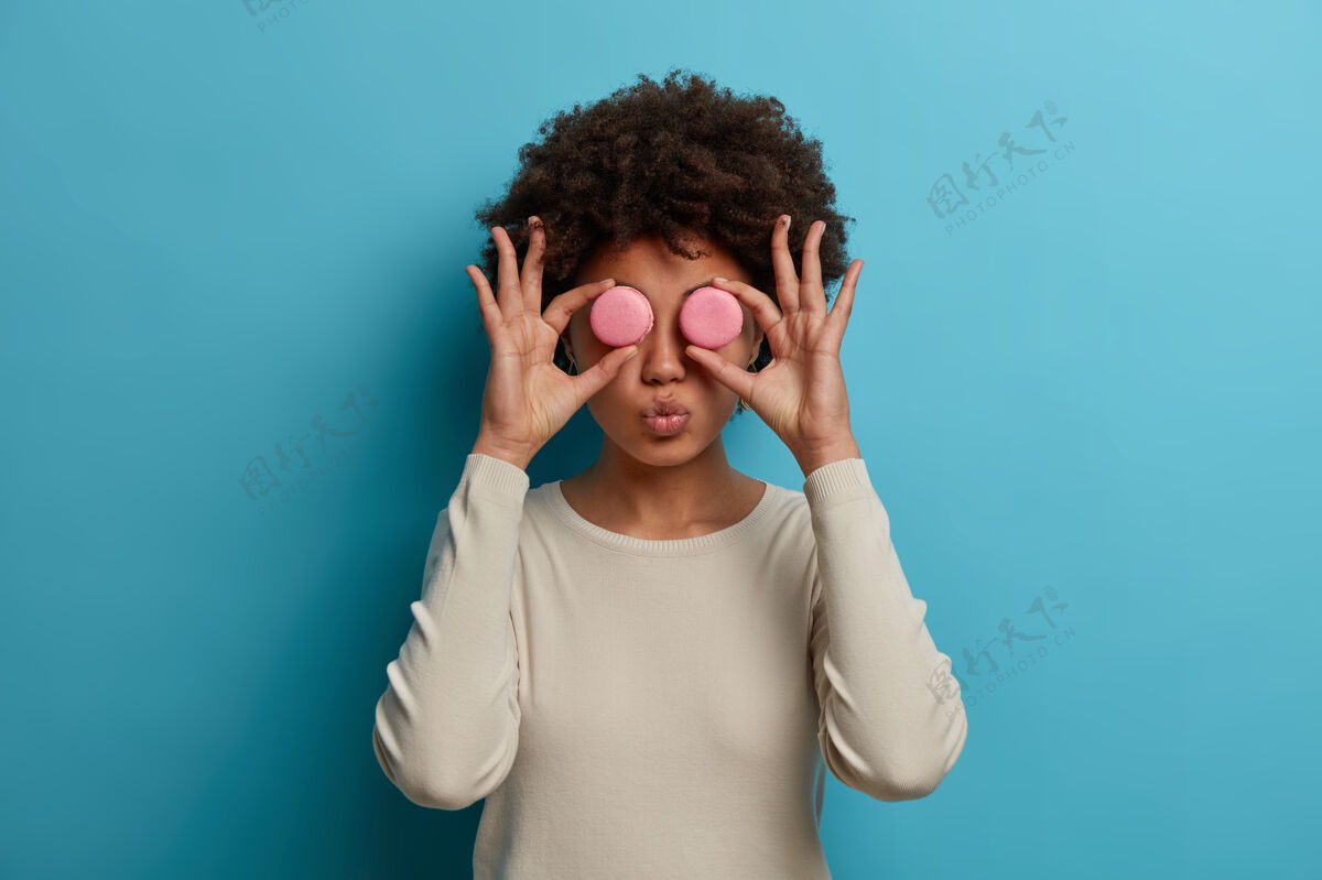 情绪有趣的女人甜食使两个粉红色的杏仁饼眼镜 保持嘴唇圆 喜欢吃高热量的法国甜点 高兴打破饮食习惯 有乐趣隔离在蓝墙垃圾食品的概念成人享受女人