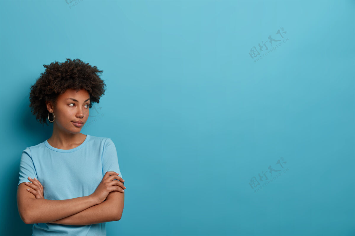 休闲体贴女人的照片有健康的皮肤 双手交叉放在胸前 注意力集中在一边 穿着休闲t恤 隔离在蓝色的墙上 空白处为您提供信息 制定计划个人女性卷发