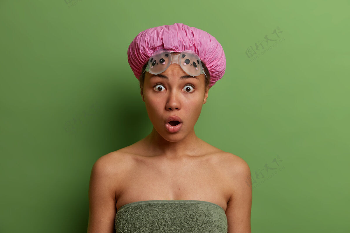 过程惊呆了的女性睁大眼睛和嘴巴盯着 对什么东西有震惊反应 戴着浴帽 护目镜和浴巾 有卫生程序 隔离在绿色的墙上女士害怕外观