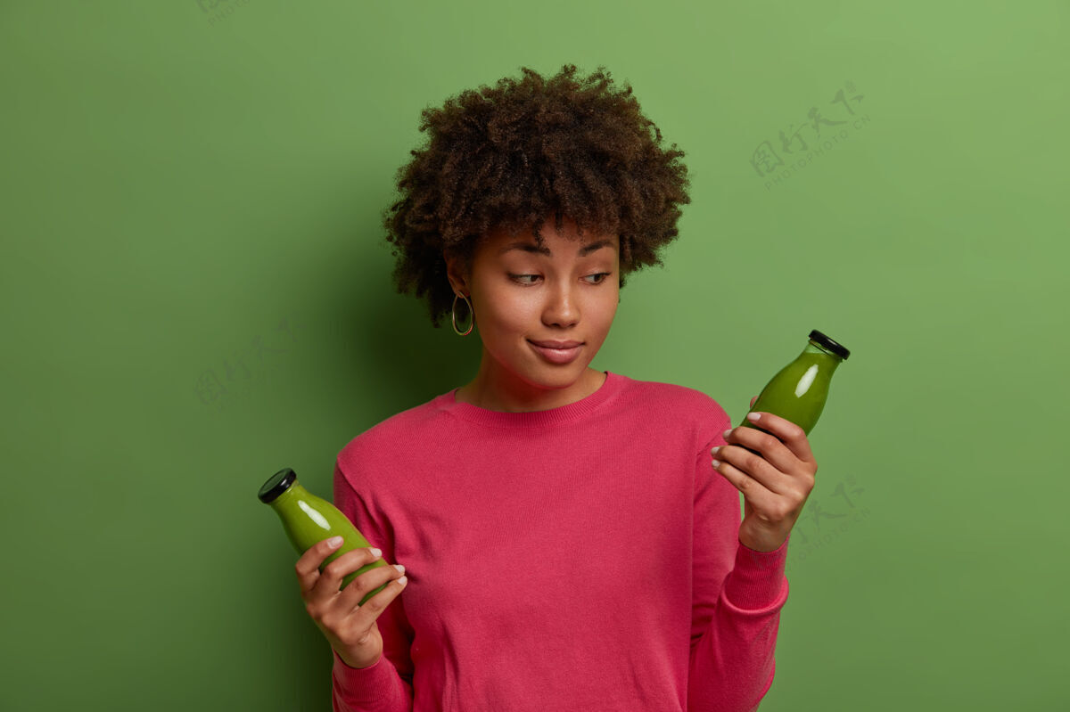 新鲜犹豫不决的黑发女人看着玻璃瓶中的排毒绿色冰沙 喝着健康的蔬菜饮料 过着健康的生活方式和适当的营养 吃着富含维生素的素食非洲女士高兴