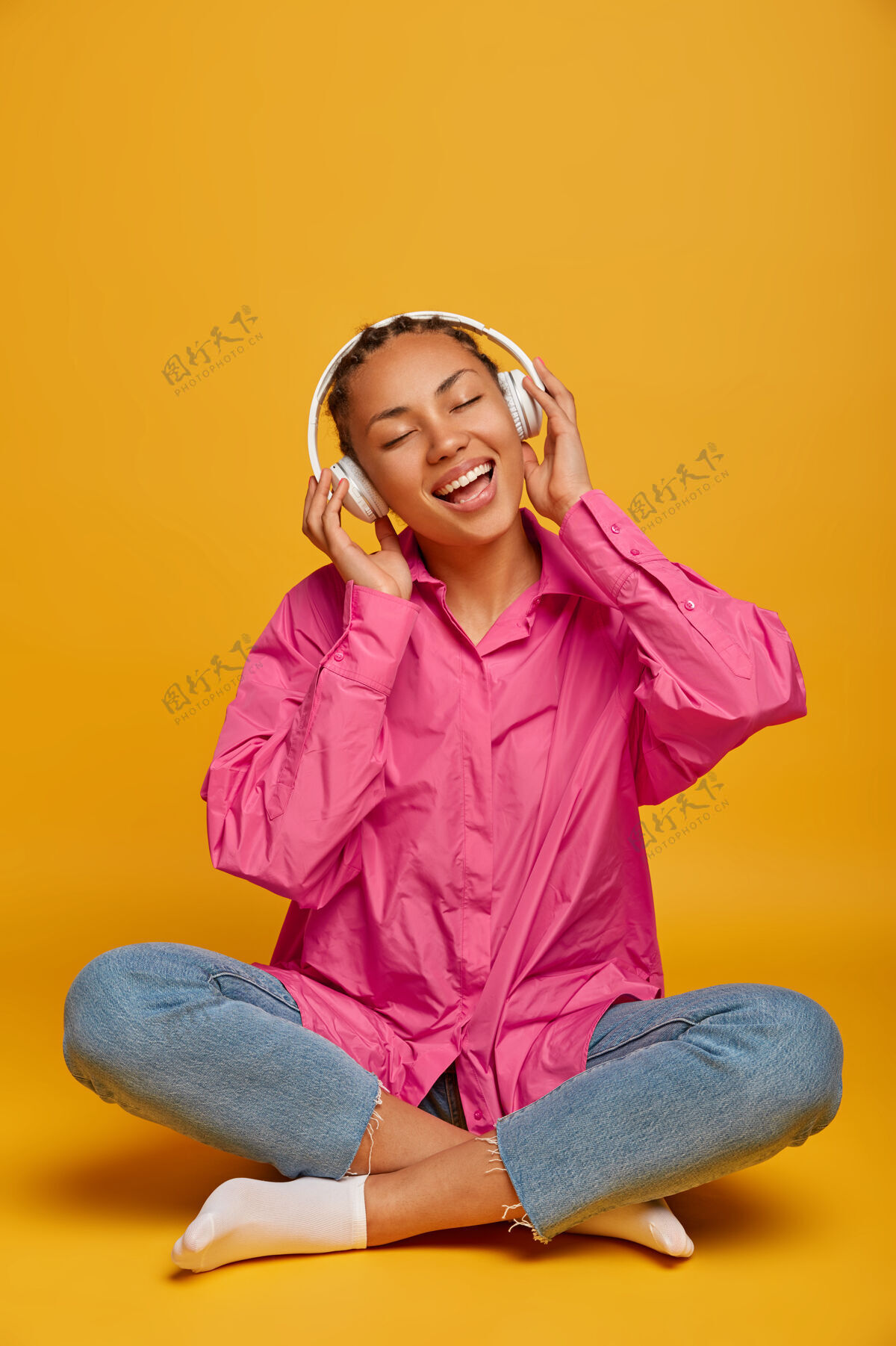 年轻年轻开朗的民族妇女在地板上欣赏音乐 盘腿坐着 穿着粉色衬衫 牛仔裤和袜子 大声地听音乐 隔离在黄色的墙上 上面空无一人请心情腿