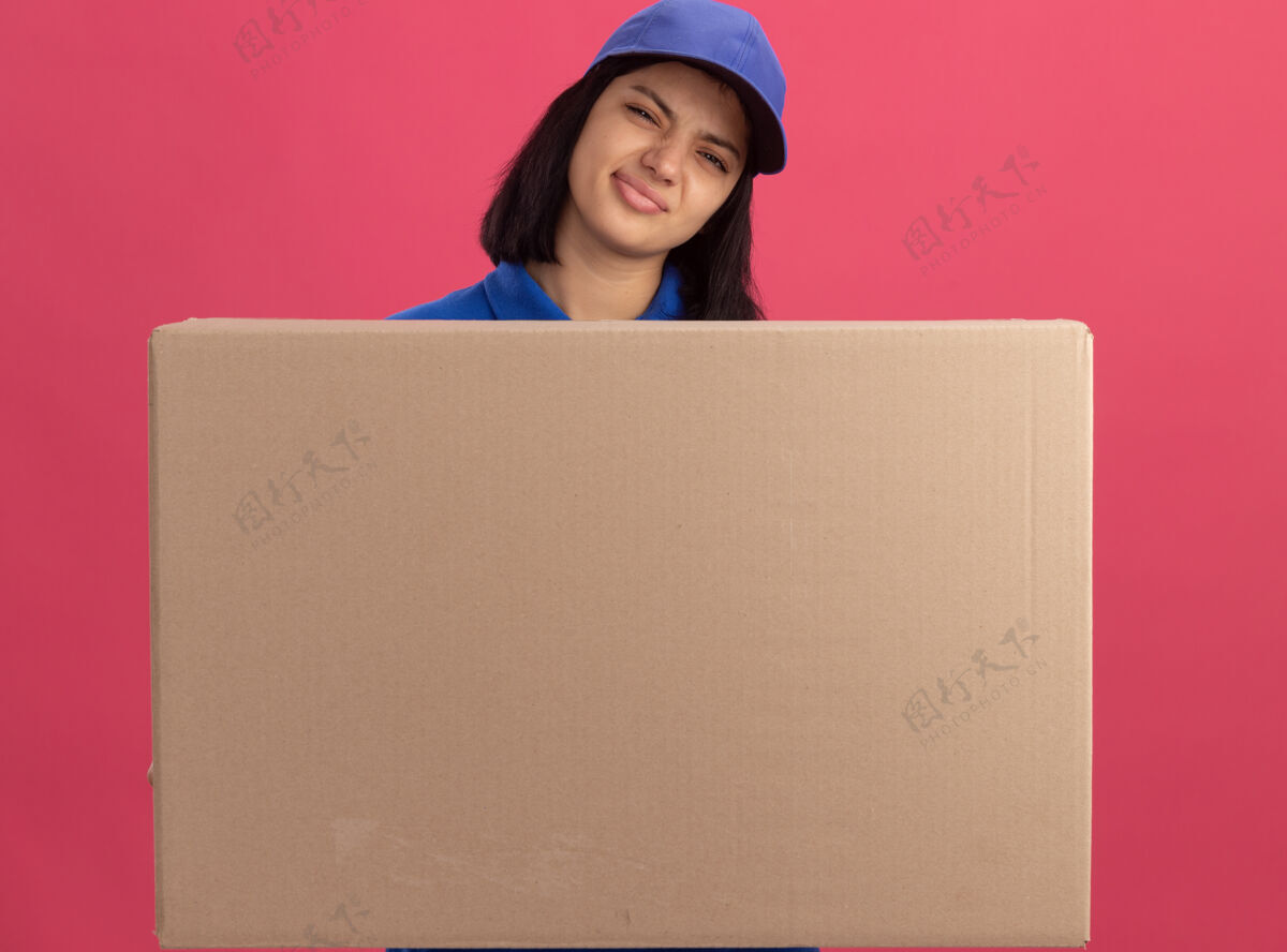使身穿蓝色制服 戴着帽子的年轻送货员站在粉红色的墙上 手里拿着一个大纸箱 嘴角歪歪扭扭的 脸上露出失望的表情年轻表情制服