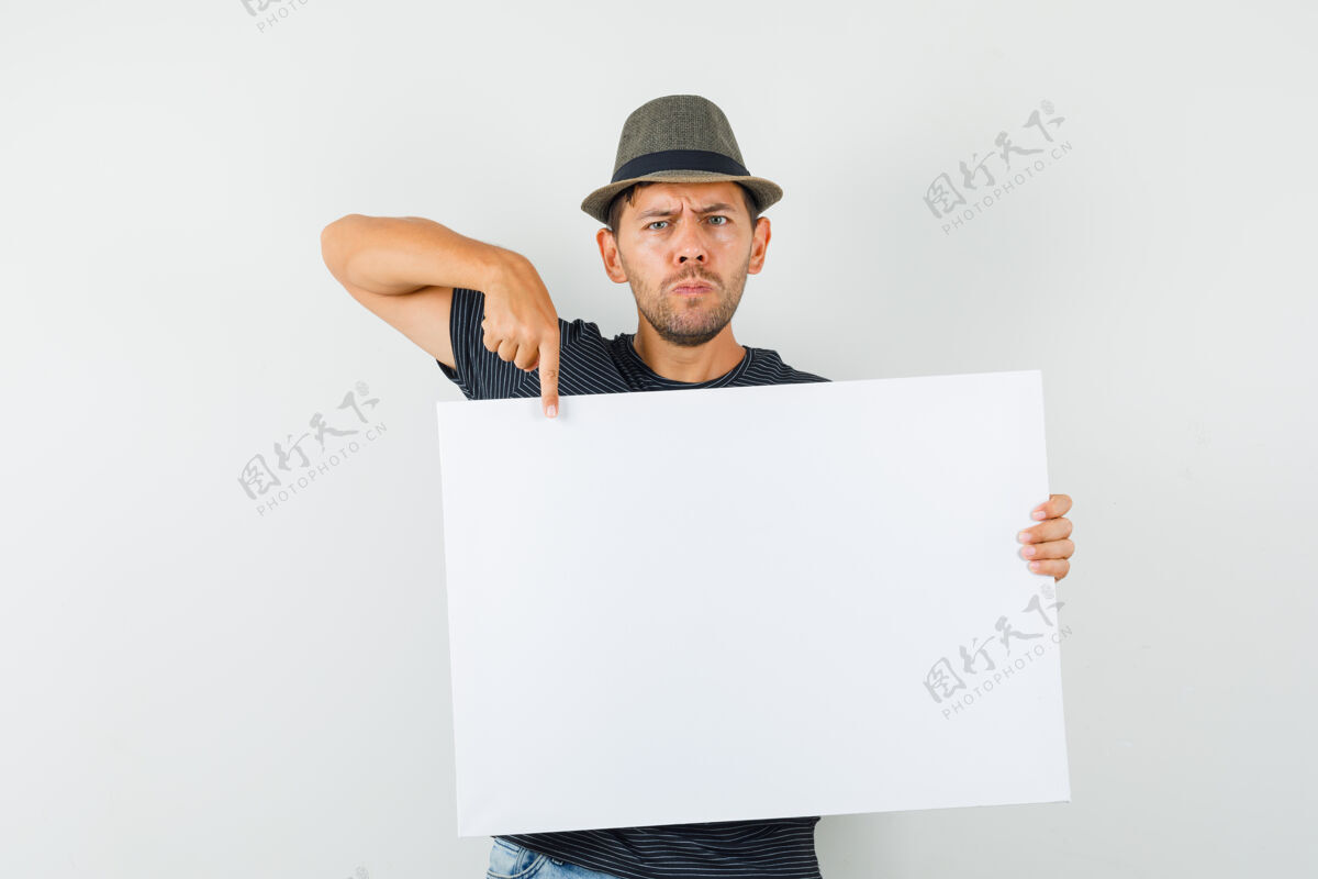 胡须年轻的男人指着一块空白的帆布 戴着t恤牛仔帽 表情严肃专业远景空白