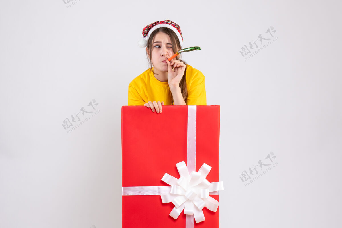 困惑的女孩前视图困惑的女孩与圣诞帽使用噪音制造站在大圣诞礼物后面立场销售圣诞节