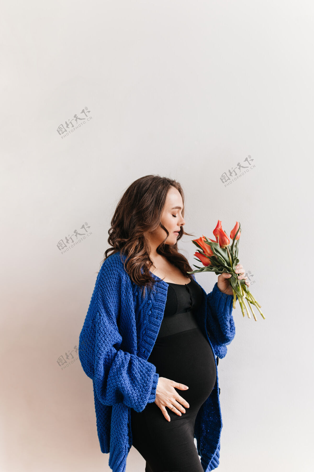 女人穿着蓝色开襟衫的快乐的卷发孕妇手持鲜花身着黑色连衣裙的迷人黑发女孩闻着白色背景上郁金香的花香白色花怀孕