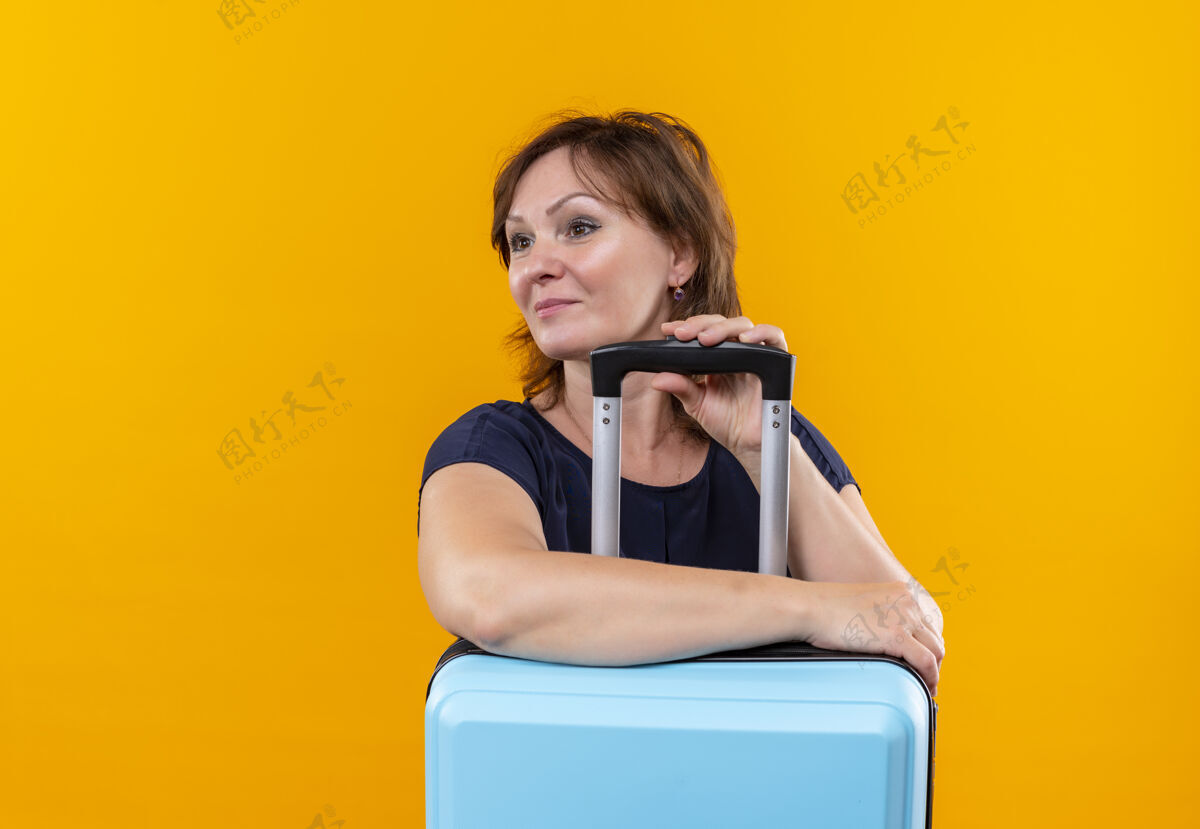 手提箱看着一旁微笑着的中年旅女把手放在箱子上孤立的黄色背景中年人旅行者壁板