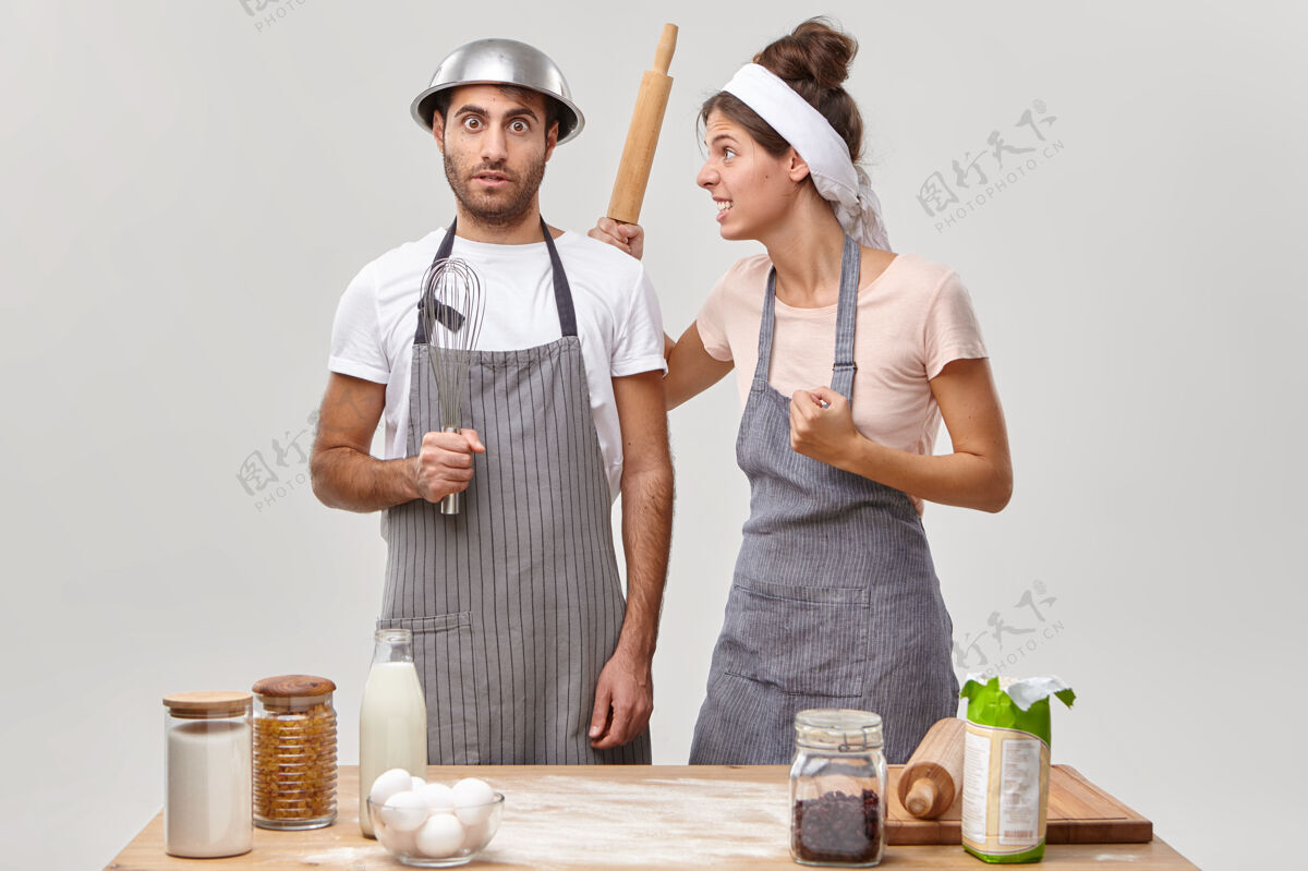 碗吵架的妻子拿着木擀面杖握紧拳头 生气地看着丈夫 让他在厨房里帮忙准备馅饼夫妻俩拿着厨具在桌边摆姿势做饭时间到了厨师头巾滚轴