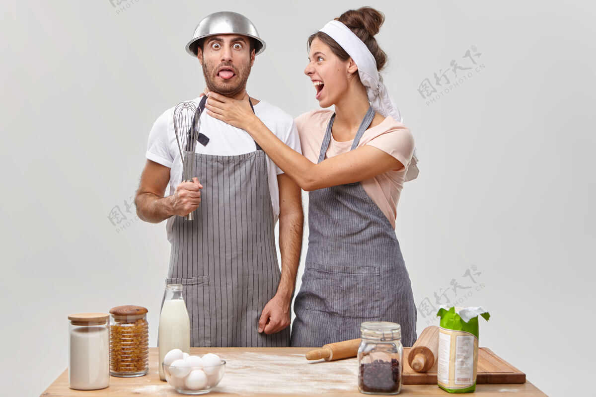 牛奶愤怒的女人把手放在丈夫的脖子上 被厨房里没有经验的厨师激怒了 一起准备好吃的东西有趣的男人头上拿着碗 手里拿着搅拌器 穿着围裙 学着做饭烹饪碗忙碌