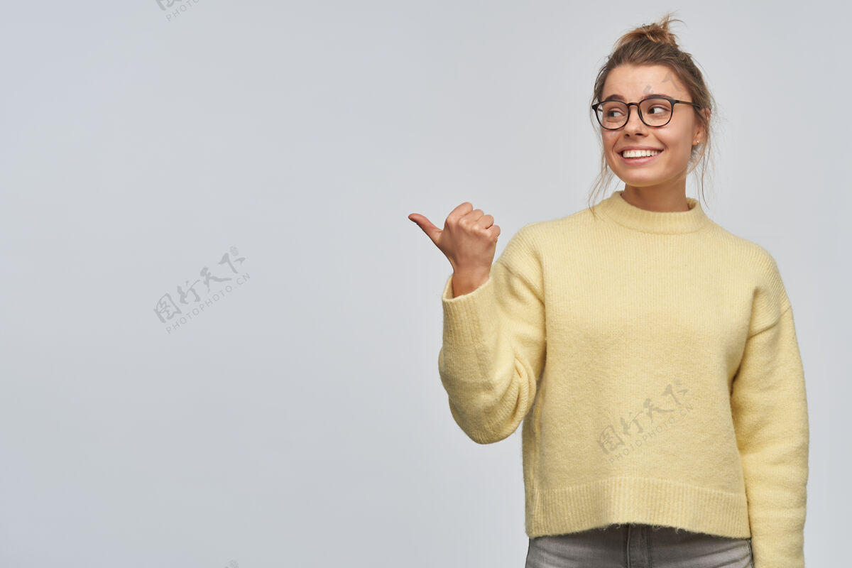 表情一幅有吸引力的成年女孩的肖像 金发束成髻 穿着黄色毛衣和眼镜 用拇指指着左边的复制空间 隔着白色的墙年轻人拇指毛衣