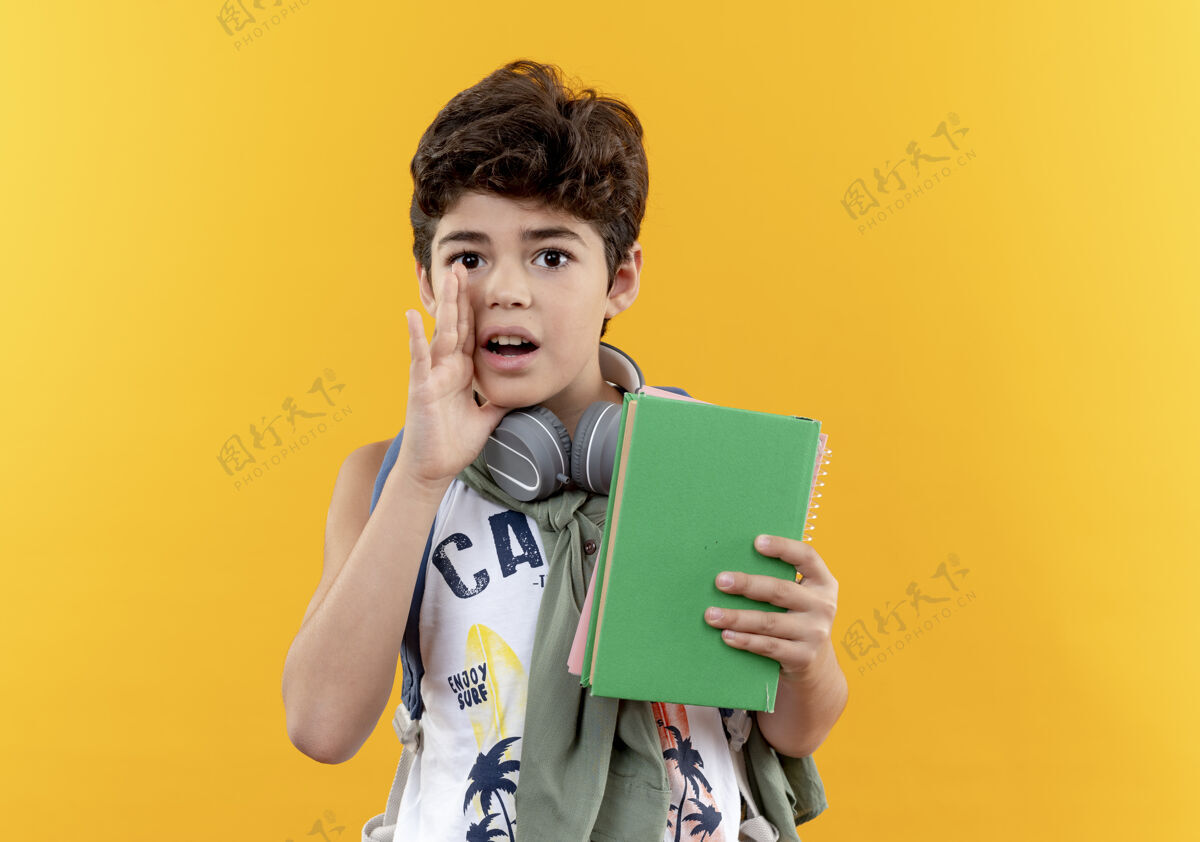 包看着摄像机 一个背着书包 戴着耳机 拿着书 在黄色背景上低语的小男孩戴男孩背