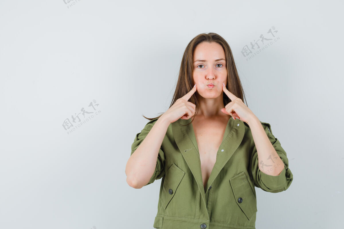黑发穿着绿色夹克的年轻女士用手指抚摸着吹得鼓鼓的脸颊吹一起女性