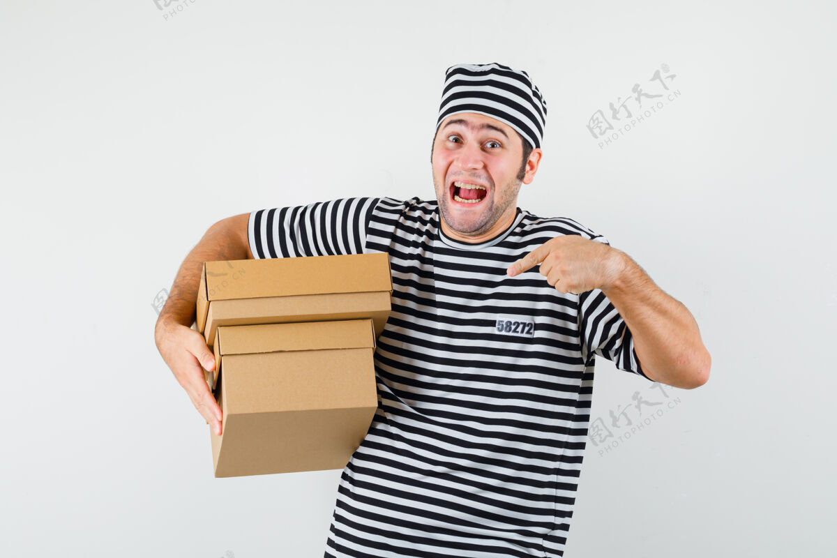 胡子年轻的男性指着穿着t恤 戴着帽子的纸板箱 看上去很开心正面图思想姿势人