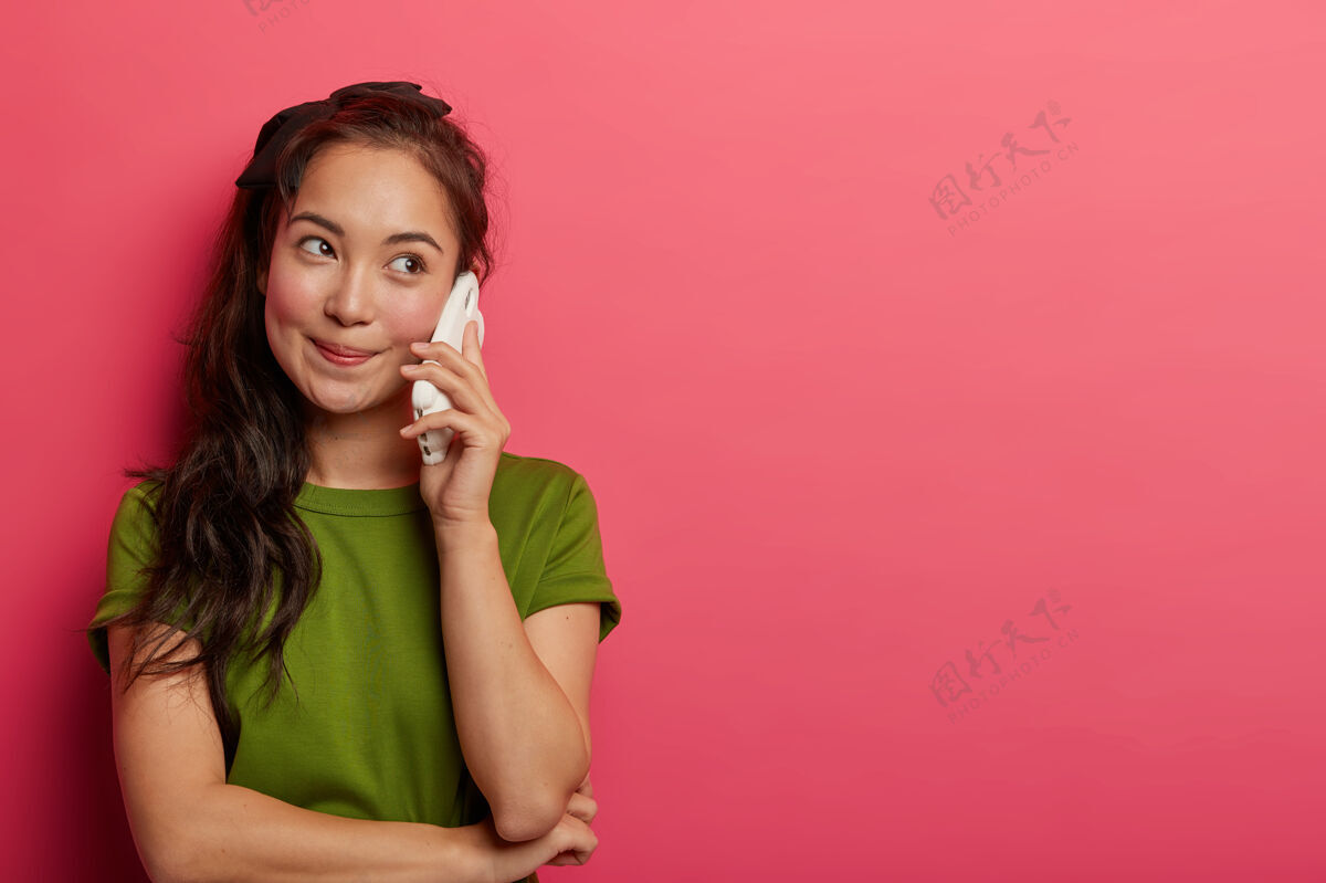 马尾辫可爱的自然黑发女孩把智能手机放在耳边 喜欢漂亮的电话交谈 看一边 穿着休闲t恤 和朋友讨论一些有趣的事情 孤立在粉红色的墙上闲话通话年轻