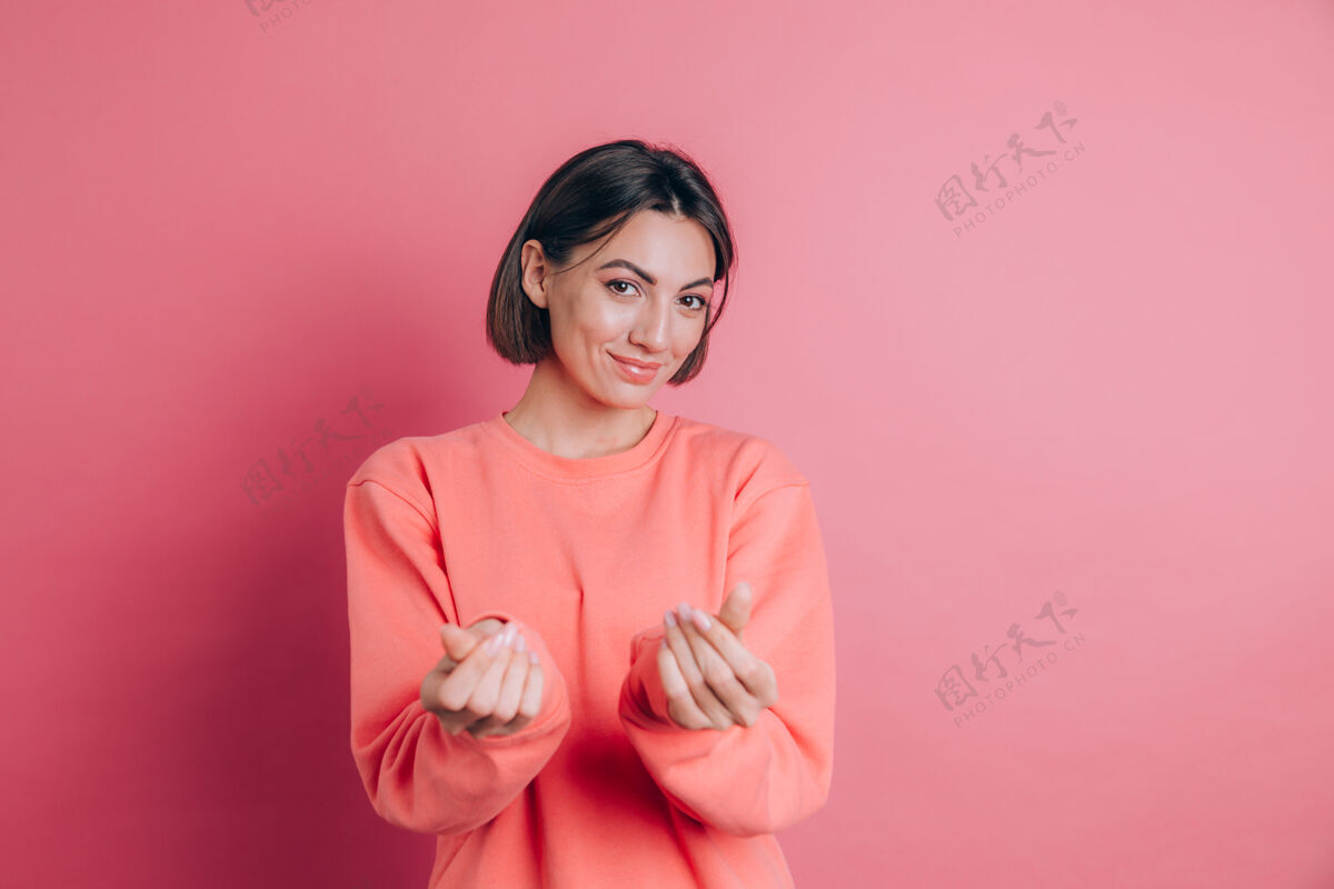 点微笑的年轻黑发女子20多岁穿着休闲站着揉手指显示现金姿态要钱粉色背景付款毛衣休闲
