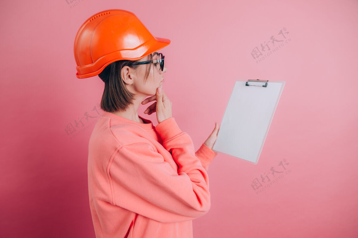 肖像漂亮体贴的女建筑工人拿着白色的空白标牌 背景是粉色的戴着建筑头盔一个保护人