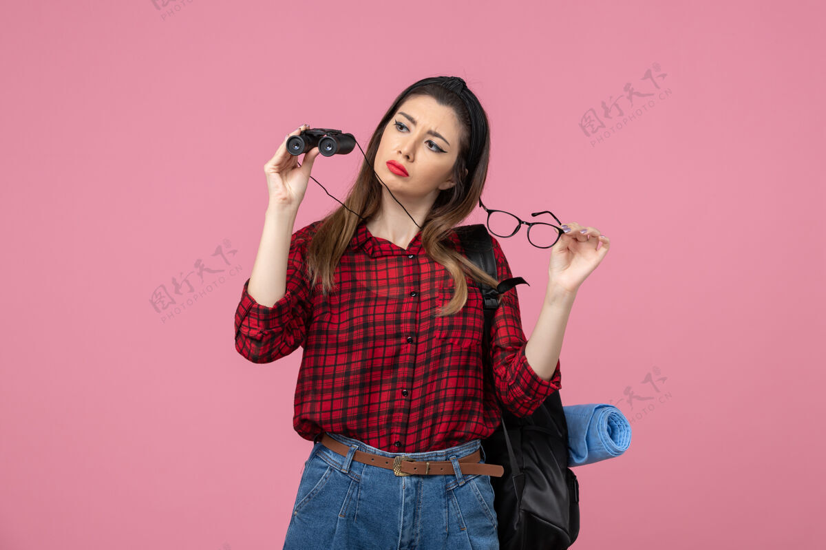 微笑正面图穿红衬衫的年轻女性用双筒望远镜在粉色背景上拍照的女性模特成人漂亮脸