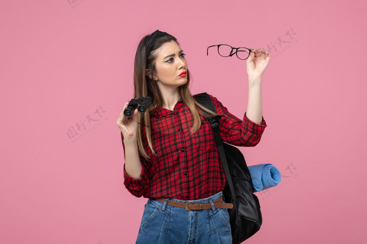 人正面图穿红衬衫的年轻女性用双筒望远镜在粉色背景上拍照的女性模特衬衫肖像年轻女性