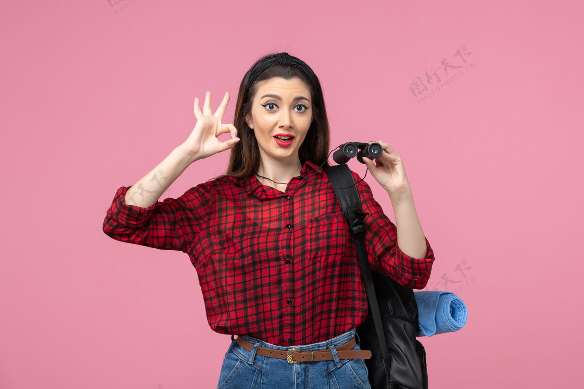脸正面图穿红色衬衫的年轻女性用双筒望远镜在粉色背景上拍摄时尚色彩的女人衬衫头发漂亮