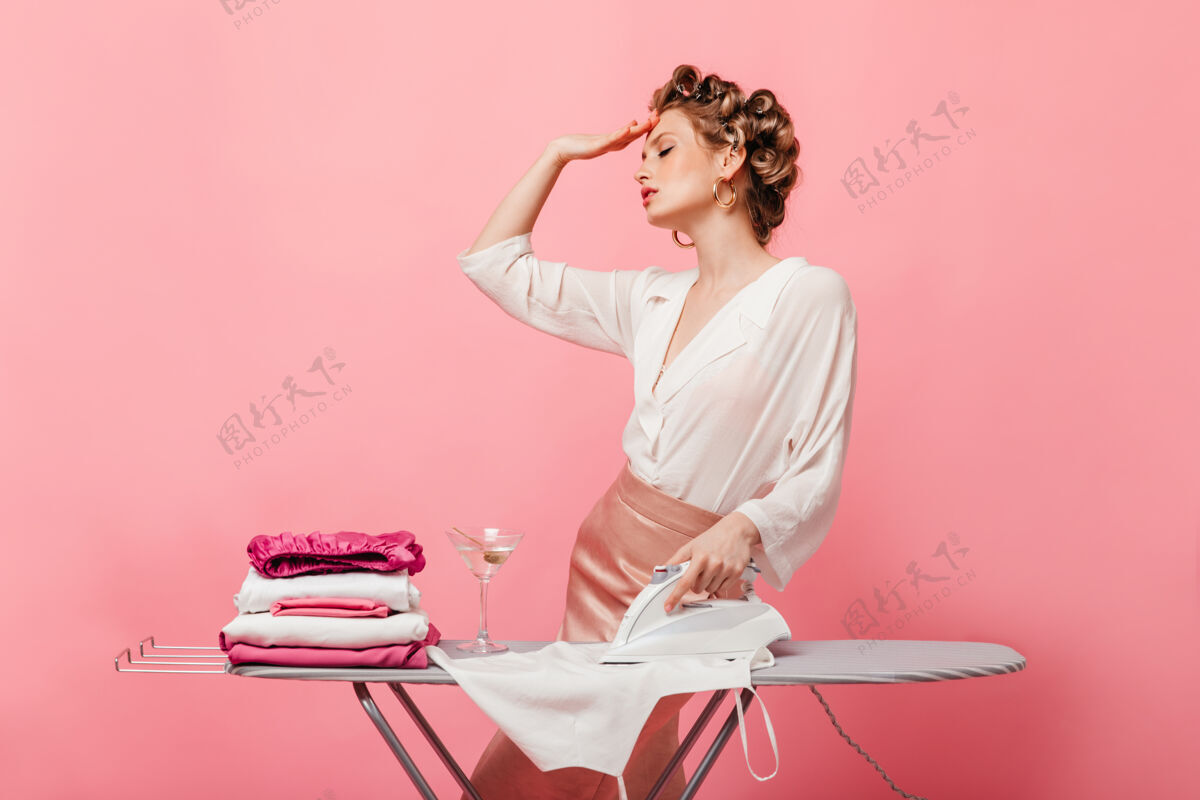 休息一个女人在熨衣服的时候疲倦地在粉红色的墙上摆姿势家庭主妇卷发成人