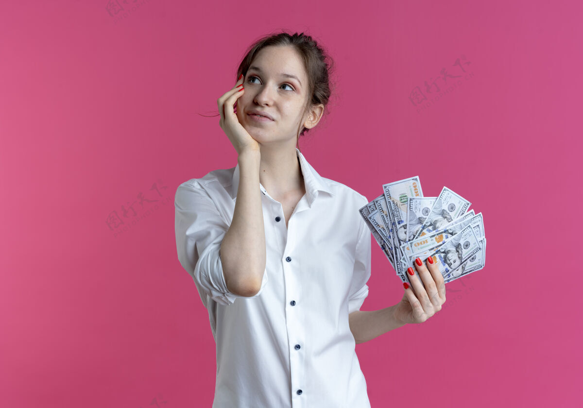 手年轻高兴的金发俄罗斯女孩把手放在脸上看一边拿着粉红色的钞票和复印空间持有空间请