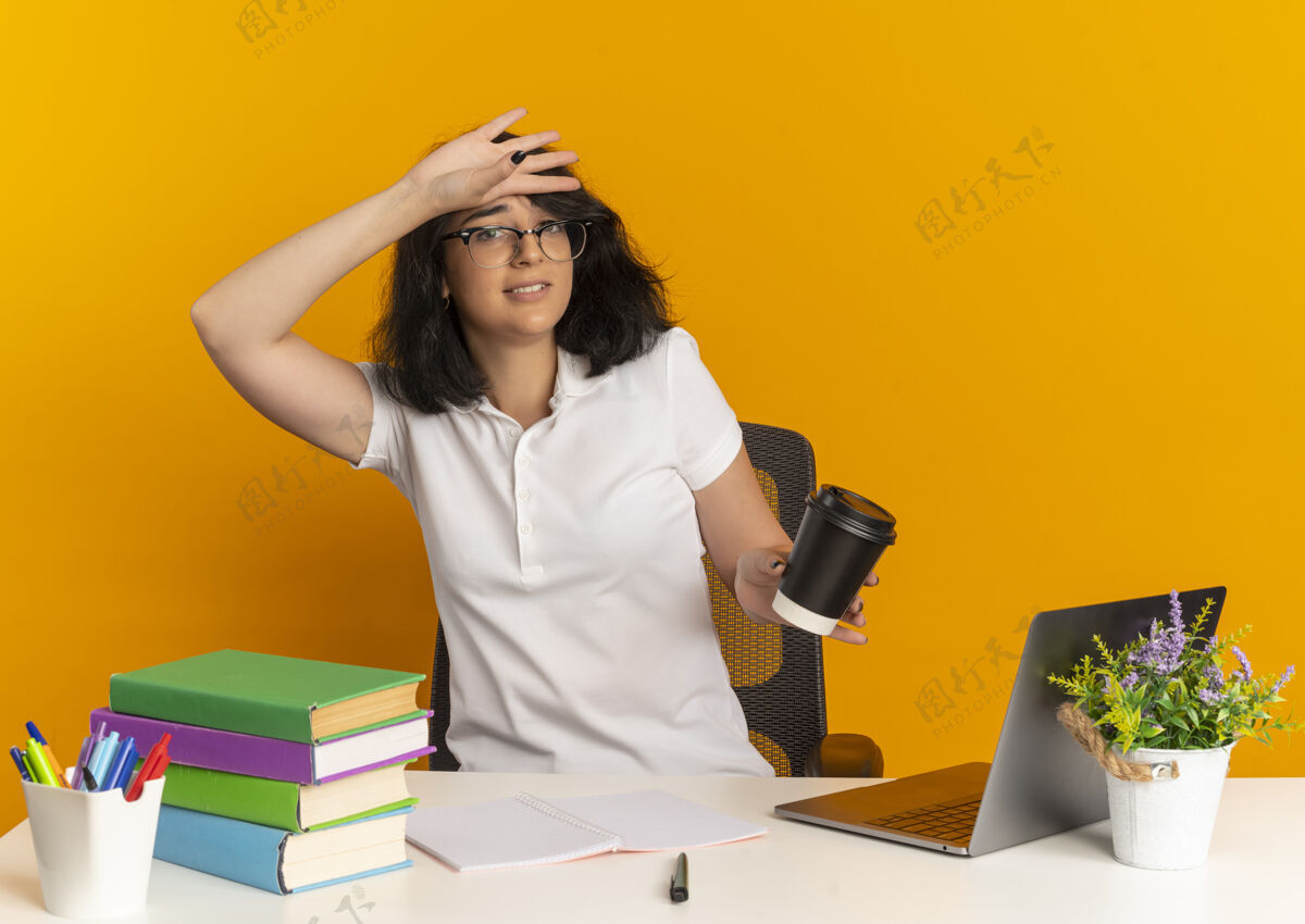 橙色年轻焦急漂亮的高加索女学生戴着眼镜坐在书桌旁 手放在额头上 拿着橘黄色的咖啡杯和复印空间复制拿着戴着