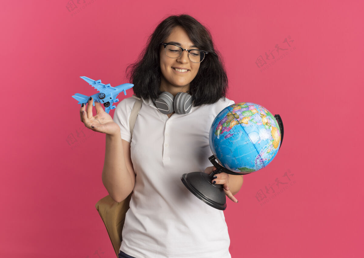 漂亮年轻可爱的白人女学生戴着耳机戴着眼镜背着袋子拿着玩具飞机和地球仪放在粉色的盒子里 还有复印空间女学生穿请