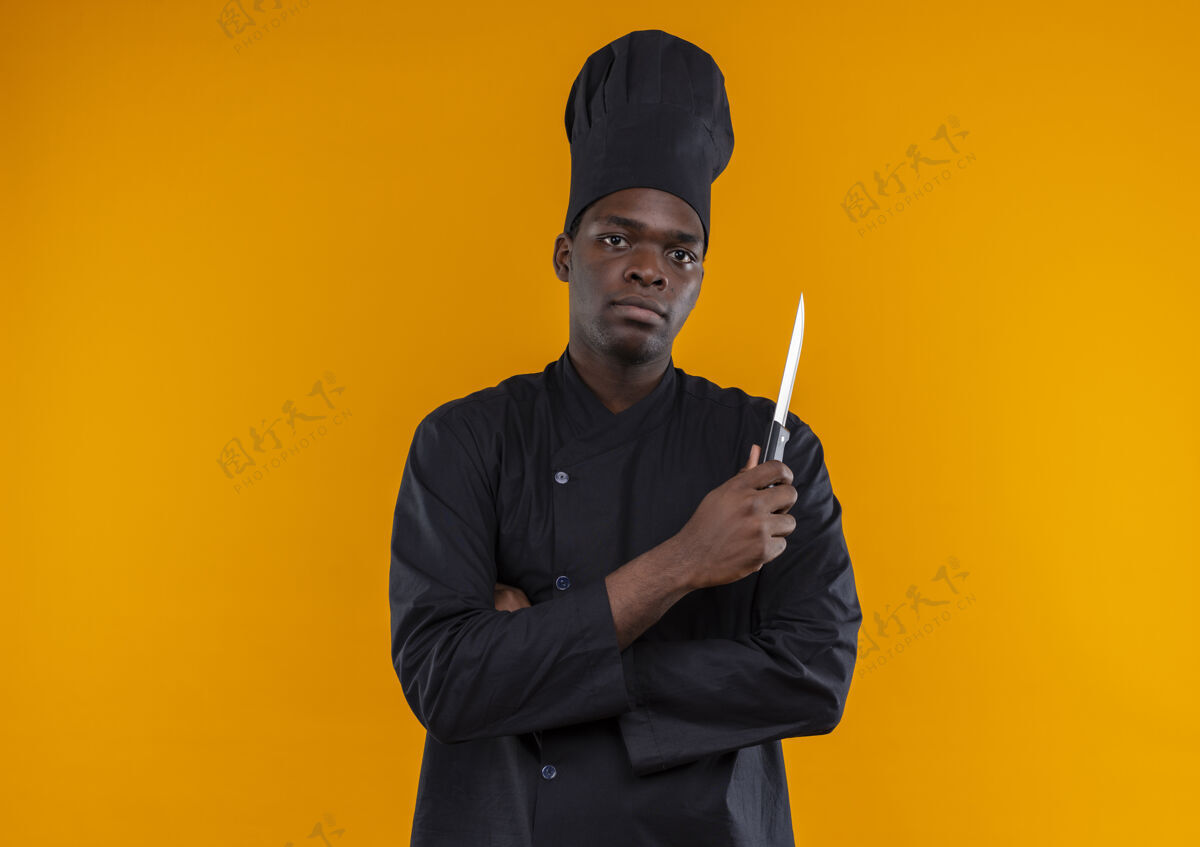 非裔美国人身着厨师制服的年轻自信的美国黑人厨师交叉双臂持刀看着橙色的相机 并留有复印空间制服交叉刀