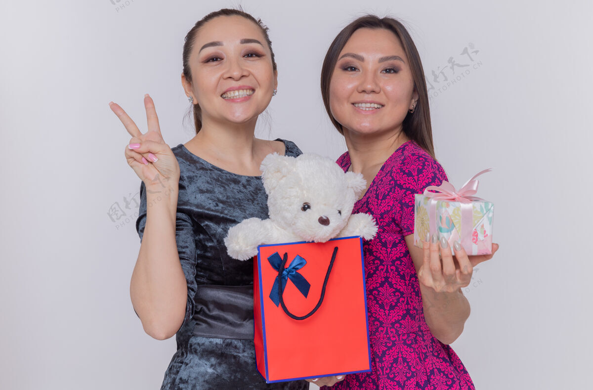 女人两位快乐的亚洲妇女拿着纸袋 手里拿着泰迪熊和礼物 喜气洋洋地微笑着 在白色的墙上立着v字标志熊泰迪纸