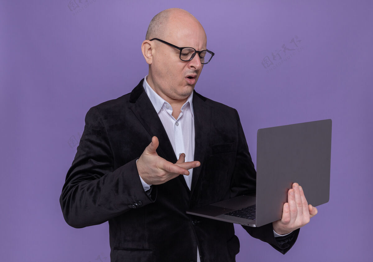 男人中年光头西装革履 戴着眼镜 拿着笔记本电脑 看着屏幕 站在紫色的墙上感到困惑和不快眼镜迷糊不高兴
