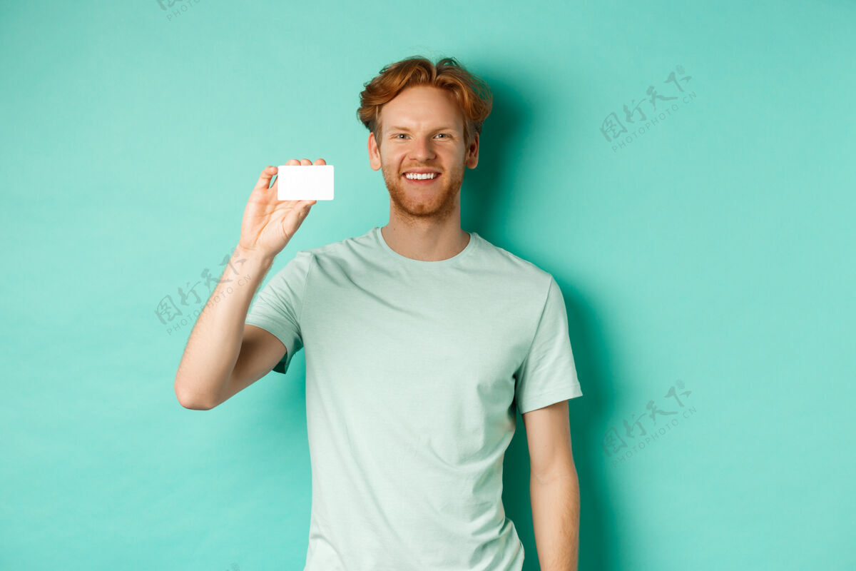 休闲购物概念帅气的红发男人穿着t恤 露出塑料信用卡 微笑着 站在绿松石背景上T恤信用银行