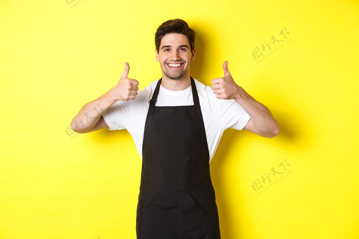 厨师穿着黑色围裙微笑的卖家竖起大拇指 表示赞同或喜欢某样东西 推荐咖啡馆或商店 黄色背景模特男人表情