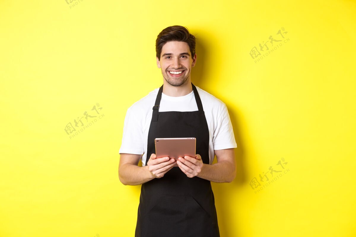 黄色身穿黑色围裙的服务员正在点菜 手里拿着数字平板电脑 友好地微笑着 站在黄色背景上围裙模特男士