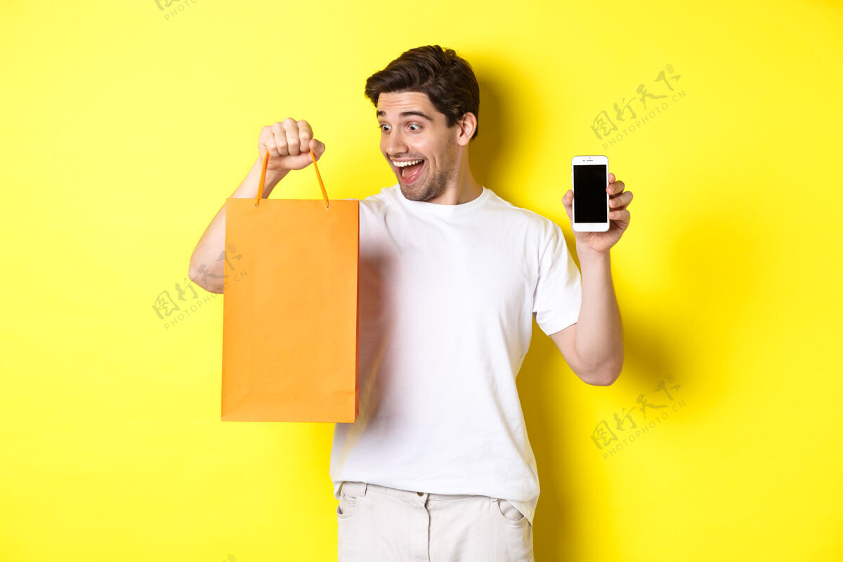 站着概念折扣 网上银行和现金返还快乐的家伙在商店买东西 看购物袋 显示手机屏幕 黄色背景表情模特男人