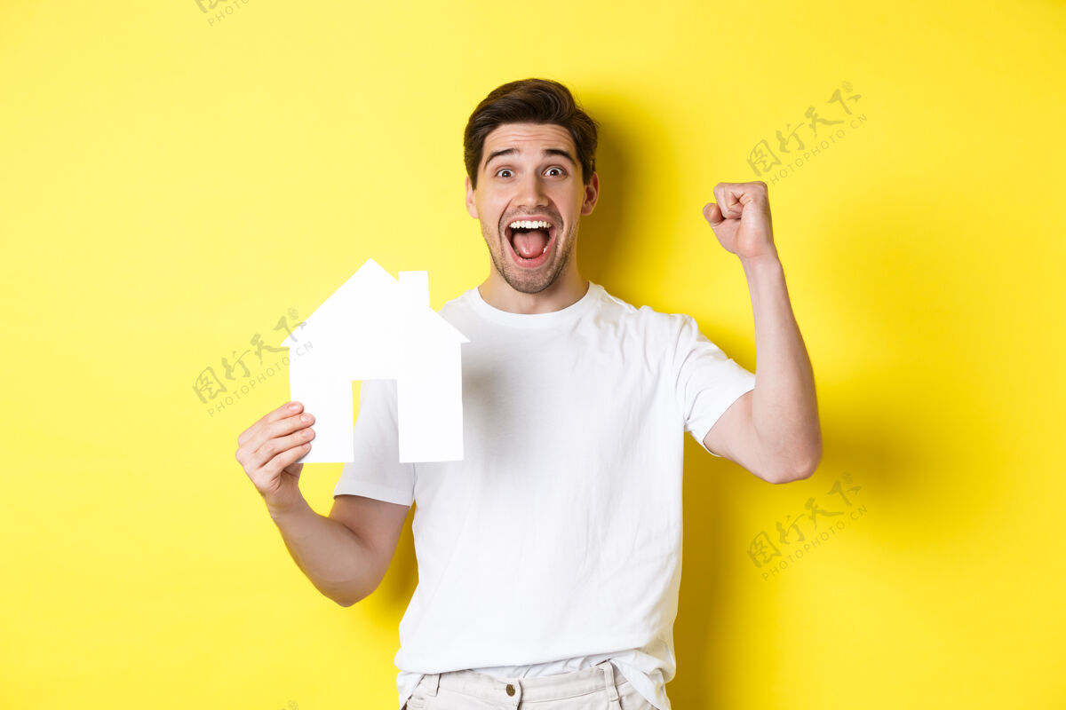时尚房地产概念开朗的男人展示纸房子模型和制作拳头泵 支付抵押贷款 黄色背景表情年轻房子