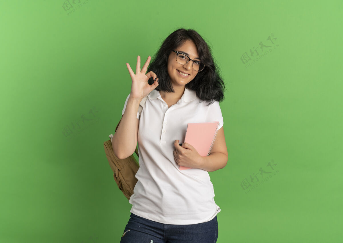手年轻的高加索女孩 面带微笑 戴着眼镜 背着书包 手势很好 手拿着笔记本 放在绿色的空白处标志空间年轻